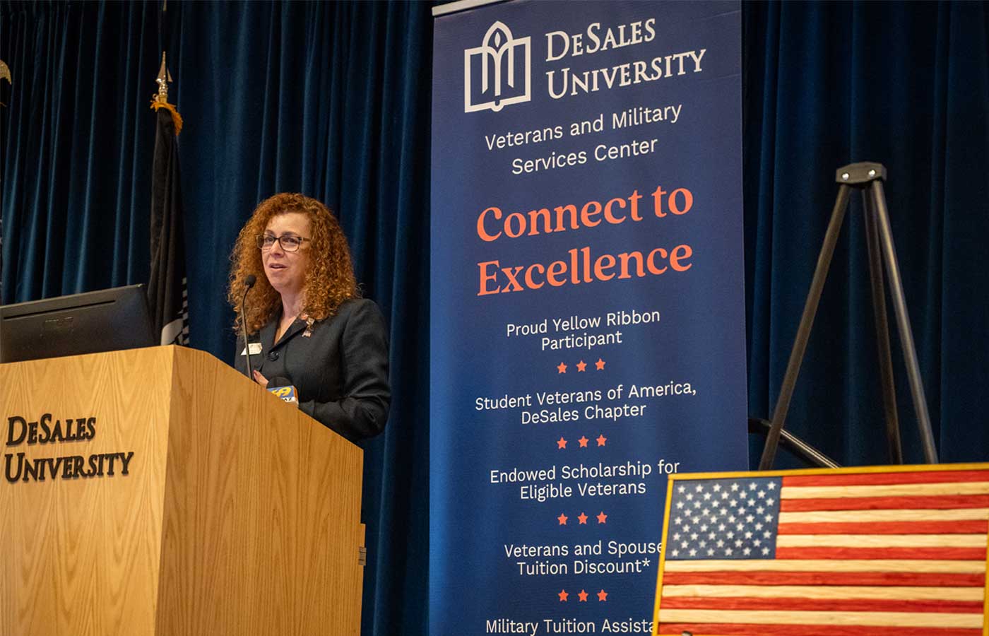 Wendy Badman, director of veterans and military services at DeSales University, speaks at a podium beside a banner highlighting the University’s Veterans and Military Services Center. An American flag display is visible in the foreground